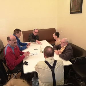 Przy stoliku kształtu prostokątnego  siedzi  6 osób. Jeden z uczestników rozdaje karty. Po lewej stronie widać innego uczestnika gry.